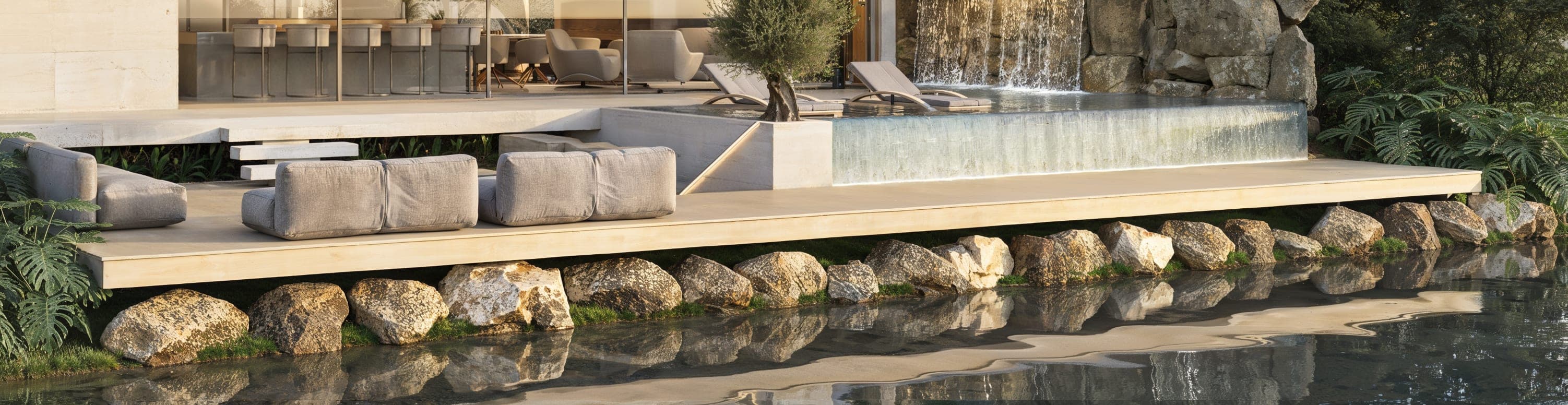 The timber deck at the water's edge: low seating and an olive tree beside the pool's overflow wall, with the boulder revetment and the lake reflecting below.